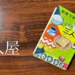 木目調のテーブルの上に置かれた原田ひ香の小説『三人屋』の表紙。カラフルな料理や飲み物のイラストが描かれており、三姉妹が営む店の雰囲気を表現している。
