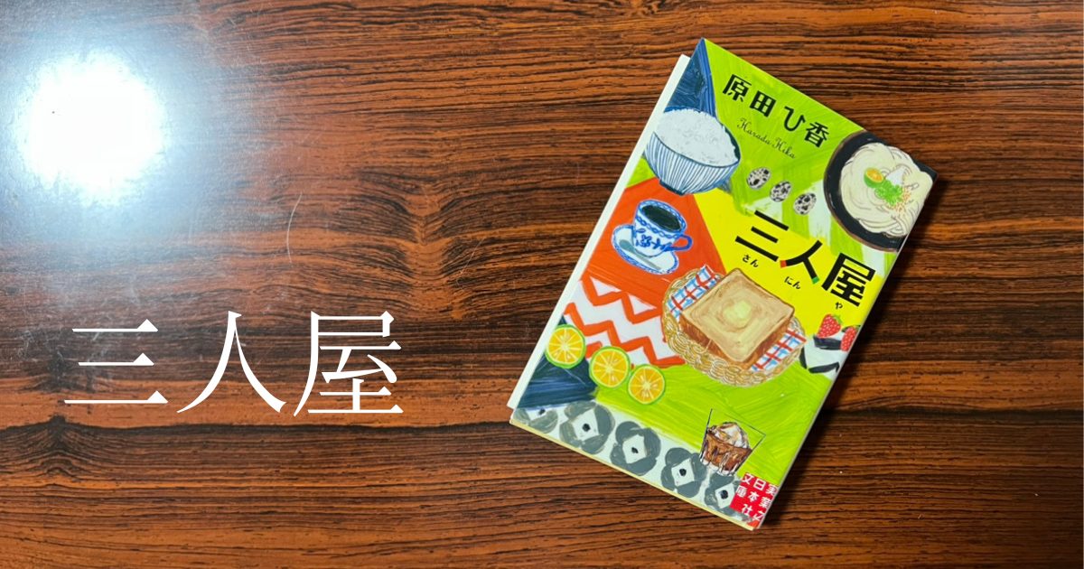 木目調のテーブルの上に置かれた原田ひ香の小説『三人屋』の表紙。カラフルな料理や飲み物のイラストが描かれており、三姉妹が営む店の雰囲気を表現している。